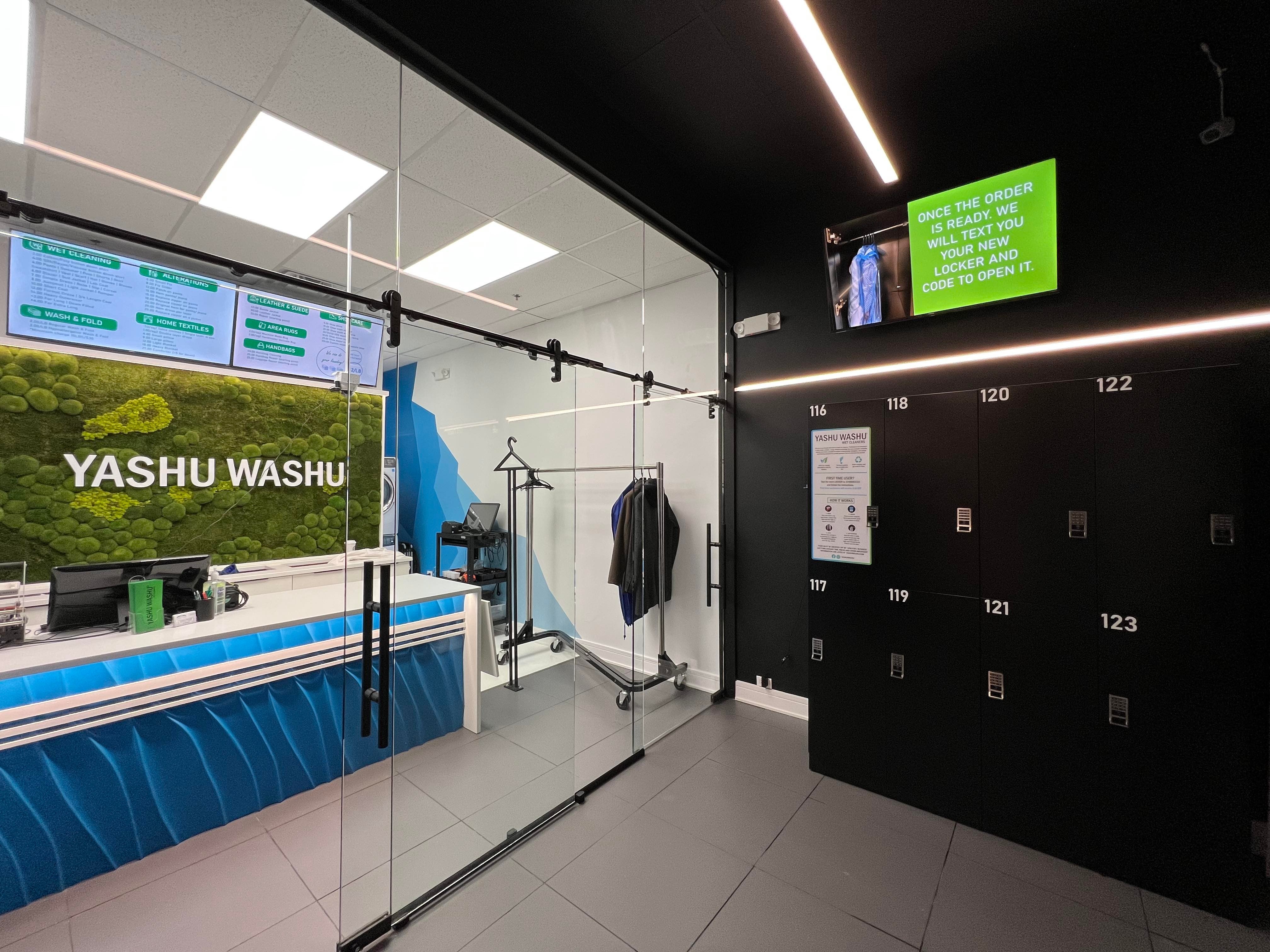 Interior of Yashu Washu retail laundry store with a bank of black numbered smart laundry lockers along the wall, automated drop-off and pickup operating 24/7